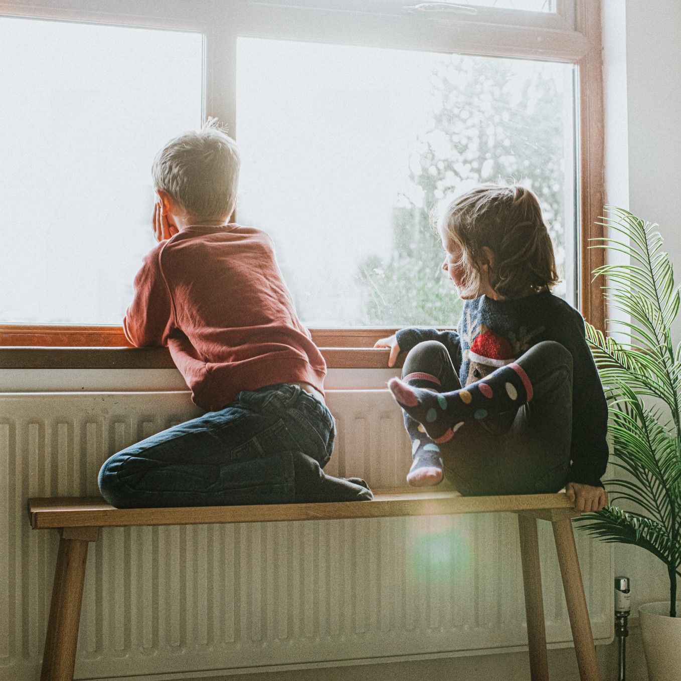 Two children are sitting on a bench looking out a window.