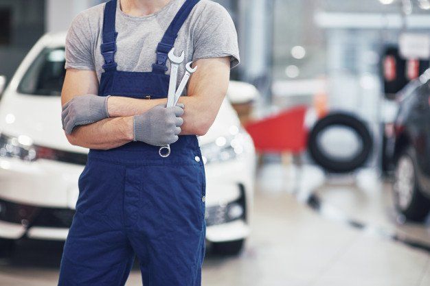 A man in overalls is holding a wrench in front of a car.