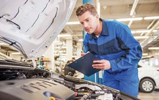 A mechanic is looking under the hood of a car while holding a clipboard.