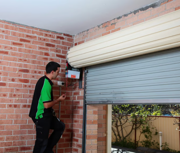 Man repairs a gray roller garage door, standing on a step stool near a brick wall.