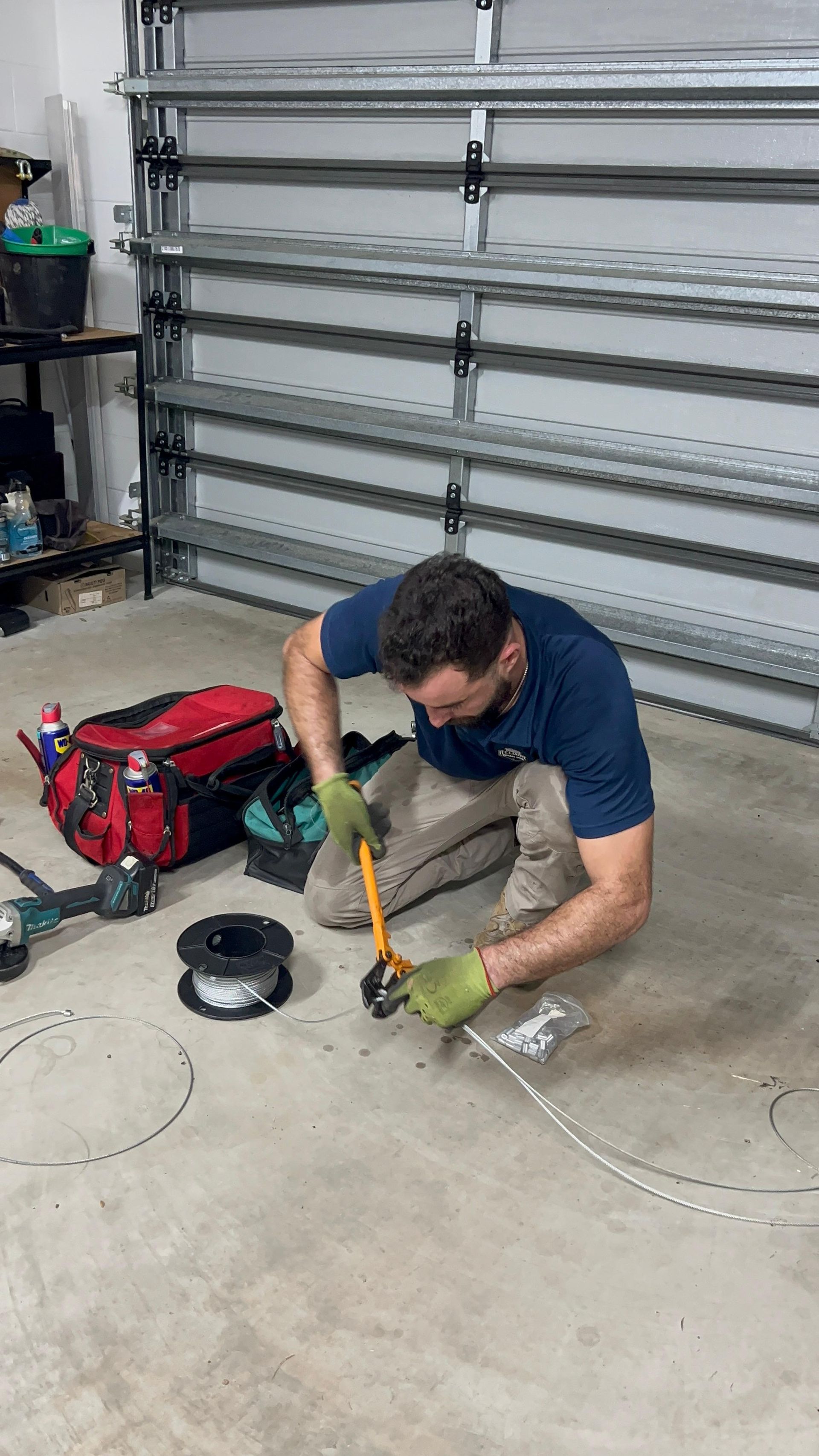 Man kneels in a garage, using a hammer on the concrete near a red toolbox and coiled cord.