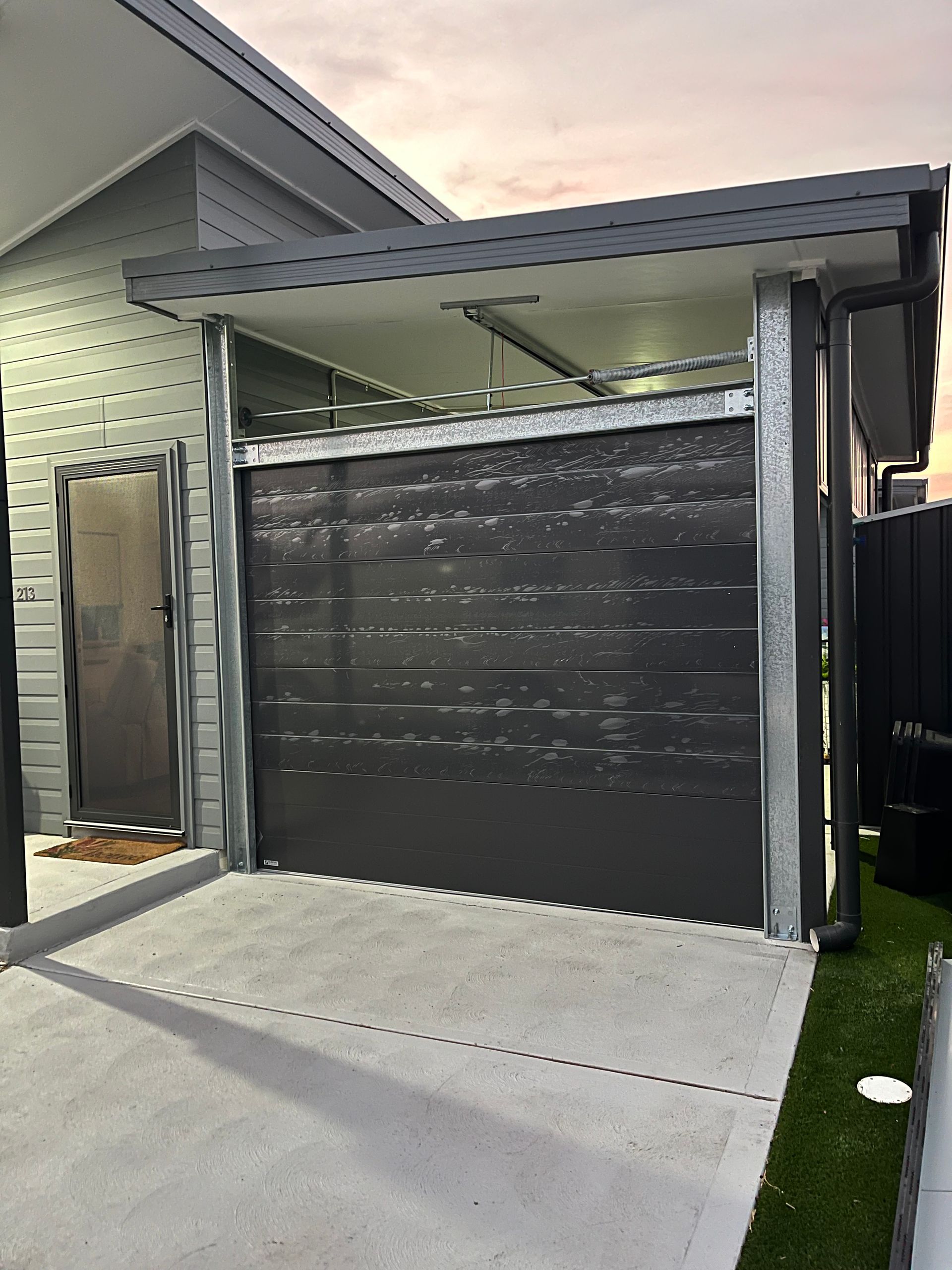 Black garage door beneath a gray metal carport beside a house, with a concrete driveway and lawn edge.