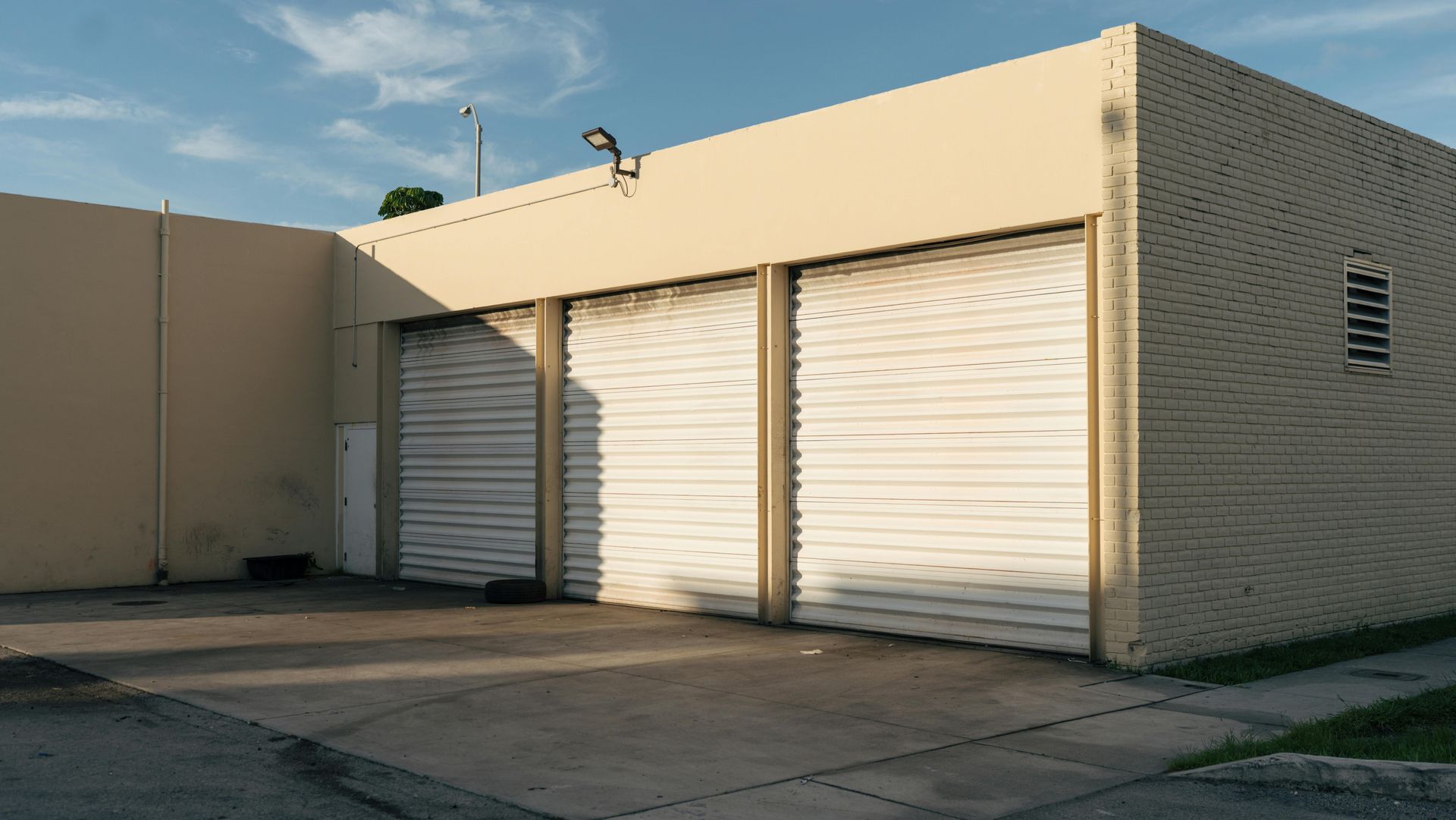 Beige brick building with three closed white roll-up garage doors in a sunny parking area