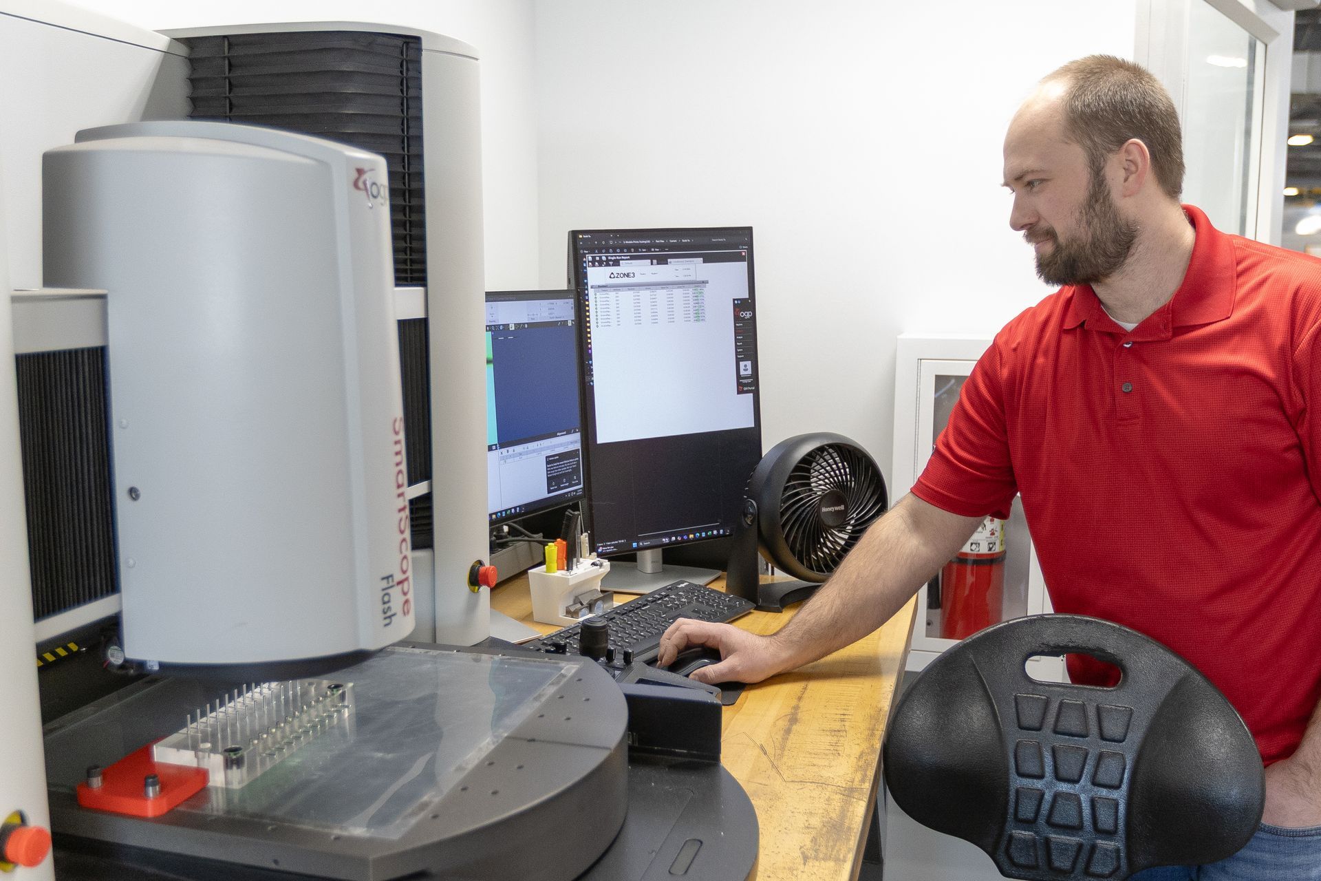Man operating a precision measuring machine in a lab. He is using a computer, with a black desk chair.