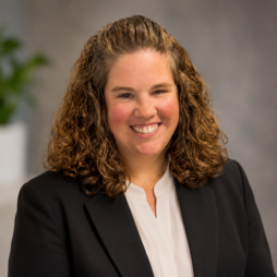 Smiling person in a black blazer and white blouse against a neutral gray background