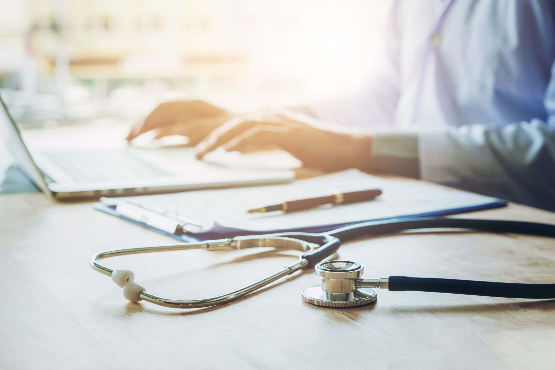 Doctor’s desk with a stethoscope, notebook, and laptop in bright light