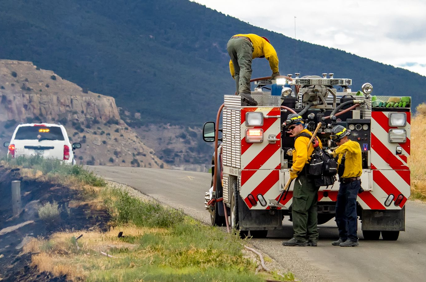 Firefighters working beside a roadside fire truck on a mountain highway, with a white car ahead.
