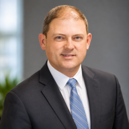 Professional headshot of a man in a dark suit and blue striped tie against a blurred office background