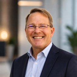 Smiling man in glasses and a navy blazer standing outdoors with a blurred building background