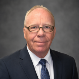 Formal headshot of a man in a dark suit, white shirt, and blue tie against a gray background