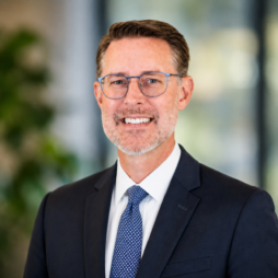 Portrait of a smiling man in a navy suit and blue tie, wearing glasses, against a blurred office background
