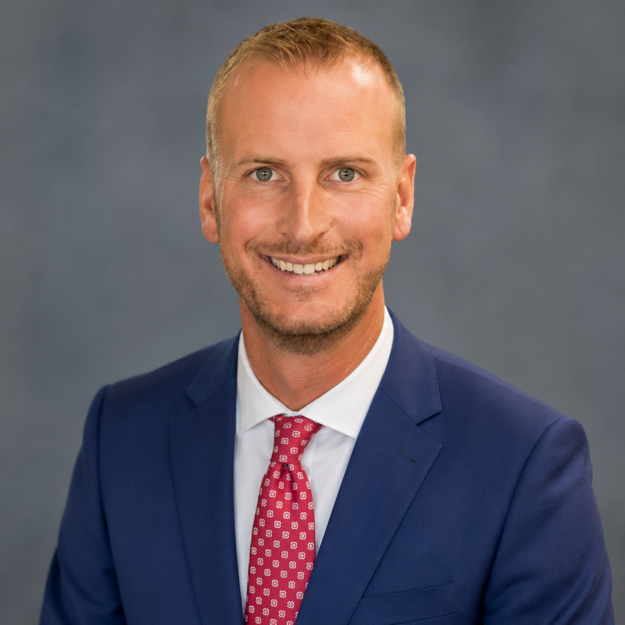 Portrait of a smiling man in a navy suit and pink tie against a gray background
