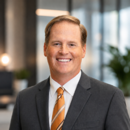 Smiling professional in a gray suit and orange striped tie, standing in a modern office.
