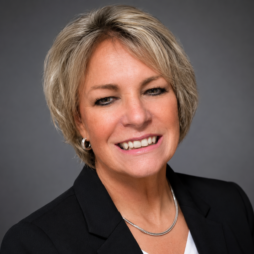 Professional headshot of a smiling person in a black blazer on a gray background