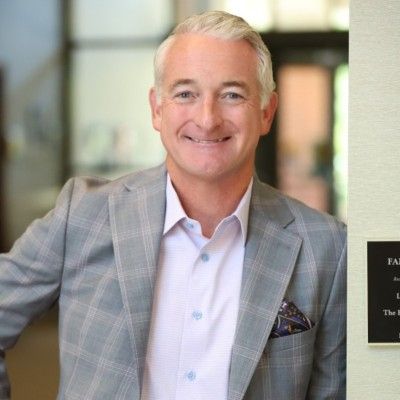Smiling man in a light gray plaid blazer standing indoors beside a wall plaque.
