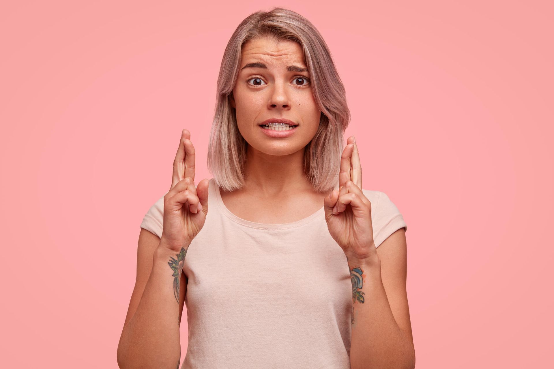 Person with raised index and middle fingers, worried expression, against a pink background