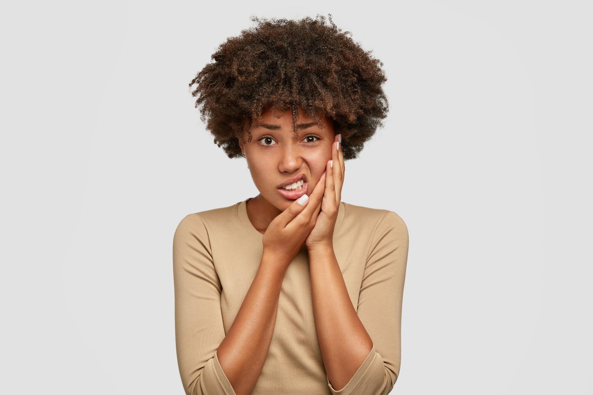 Person with curly hair touching their face, looking worried against a plain light-gray background