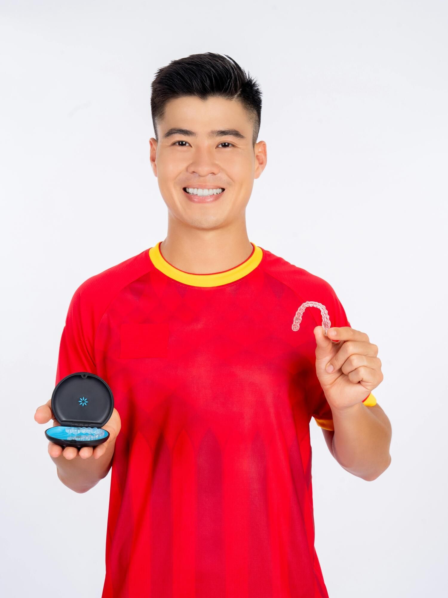 Man smiles, holding clear aligner and case against white background; wears red shirt.