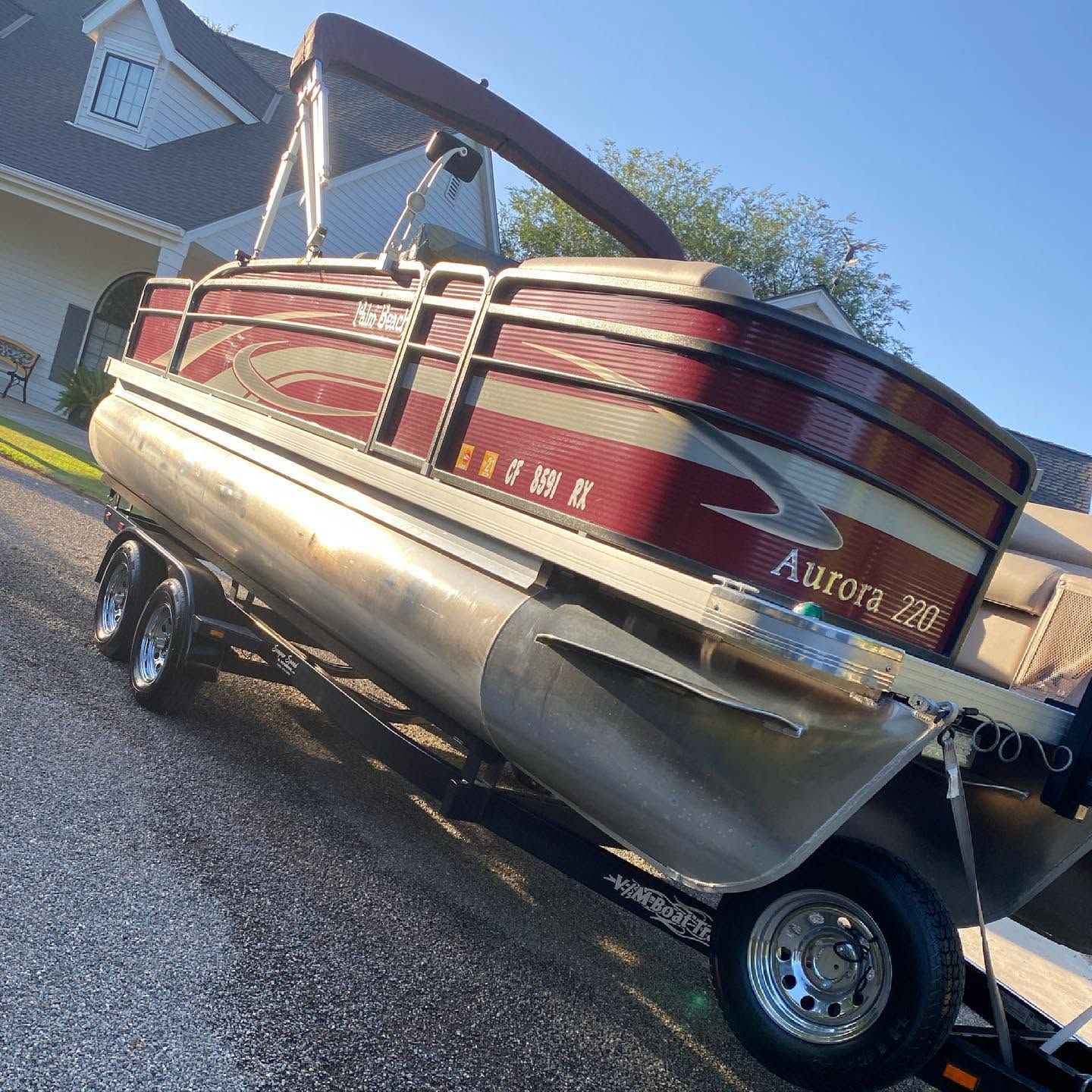 Red pontoon boat on a trailer in a driveway, with a blue house in the background.