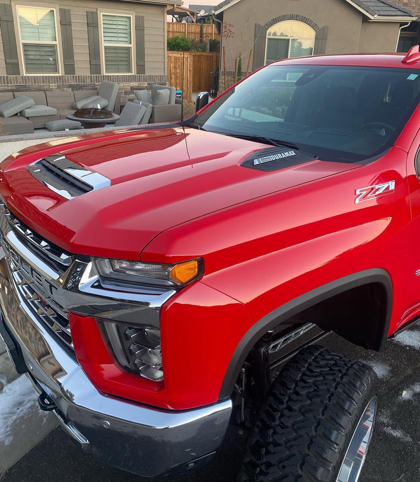 Red lifted pickup truck parked in a driveway, viewed from the front-left, with off-road tires and a chrome bumper