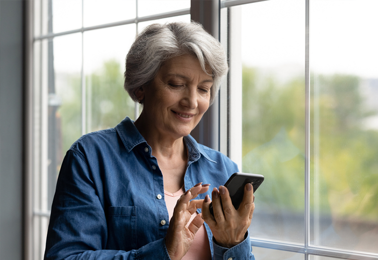 An elderly woman is standing in front of a window using a cell phone.