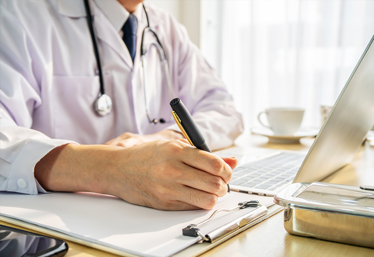 A doctor is writing on a clipboard in front of a laptop computer.