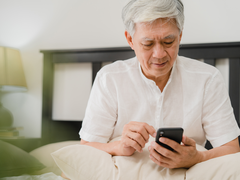 An elderly man is sitting on a bed looking at his cell phone.