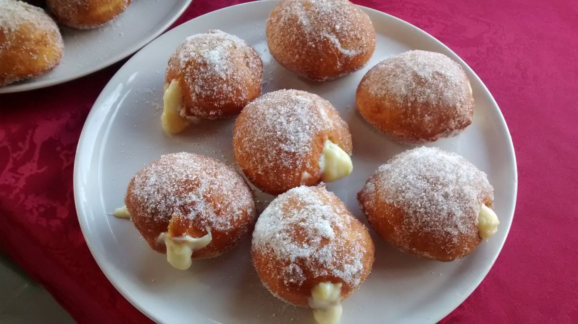 Un piatto di ciambelle ripiene di crema e cosparse di zucchero a velo, adagiato su una tovaglia rossa.