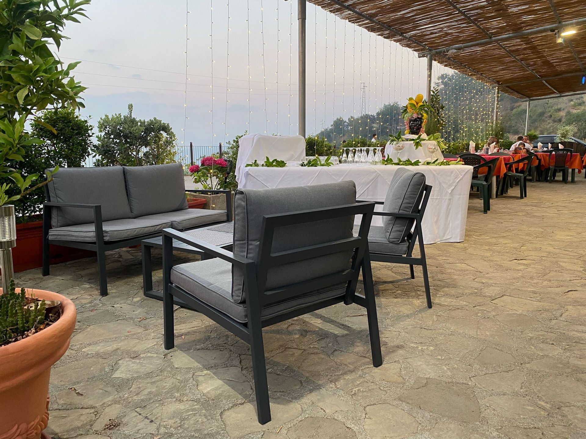 Zona pranzo all'aperto nel patio con sedie nere, tovaglie bianche, piante in vaso e luci decorative sospese.
