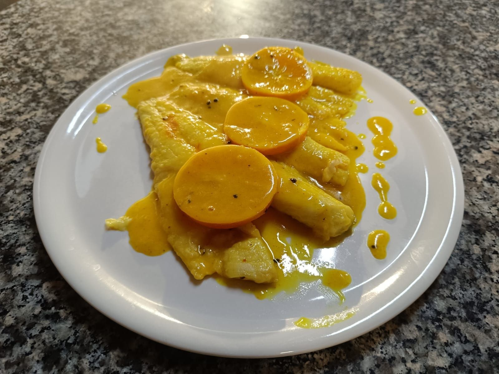 Un piatto di crêpes gialle guarnite con fette d'arancia e sciroppo su un piano di lavoro.