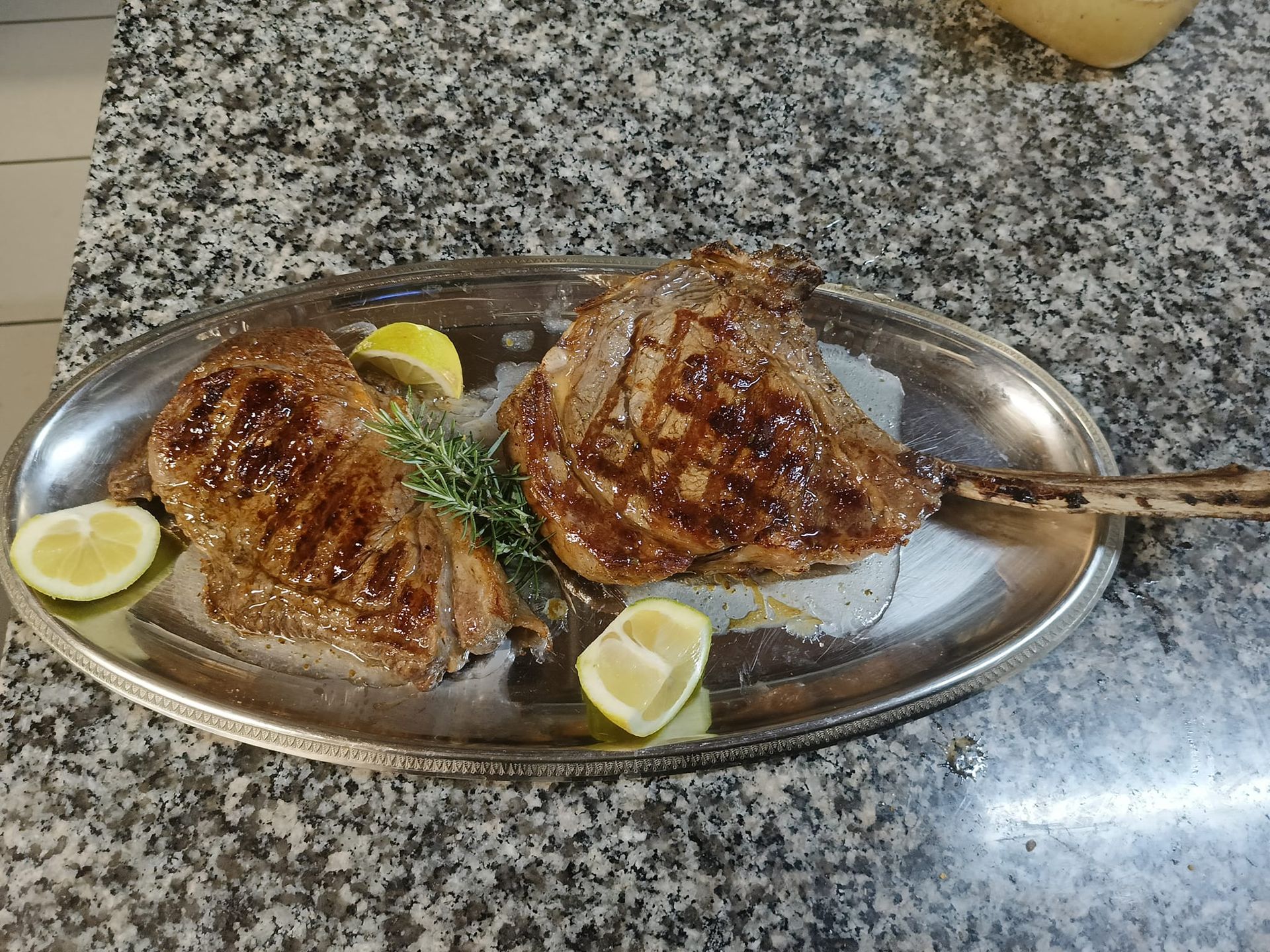 Pesce grigliato servito con spicchi di limone su un vassoio ovale di metallo su un piano di lavoro.