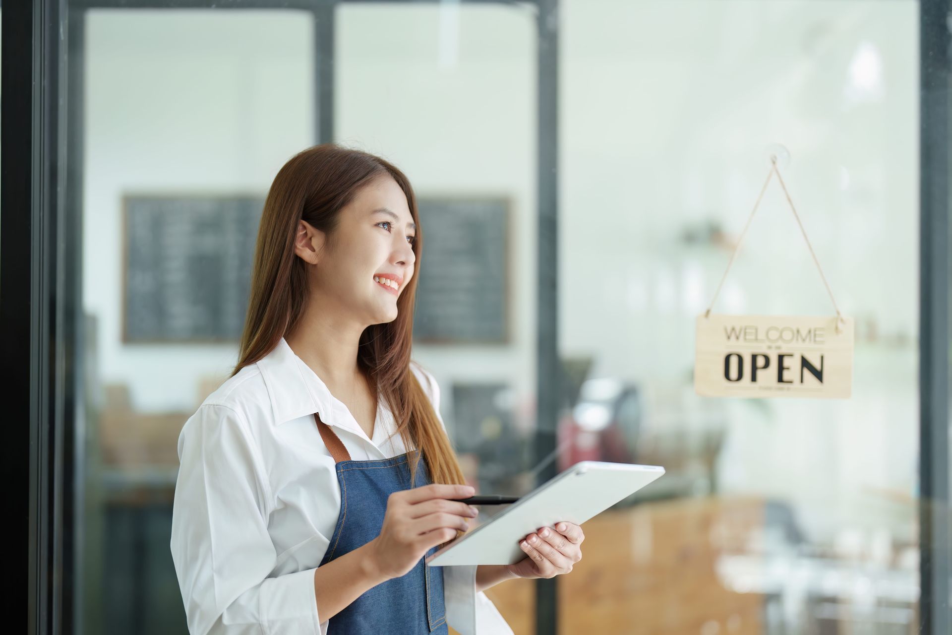 A smiling person wearing a white shirt and blue apron holds a tablet while standing near a glass door with an open sign.
