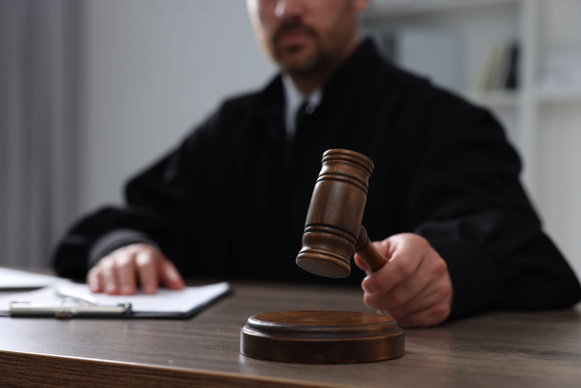A judge in a black robe holding a wooden gavel above a sounding block on a courtroom desk.