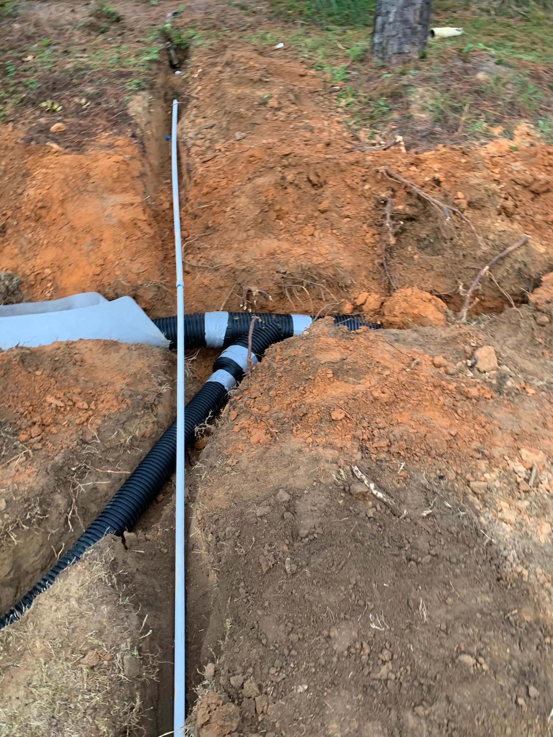 Trench with black drainage pipes and a white conduit crossing exposed orange soil outdoors