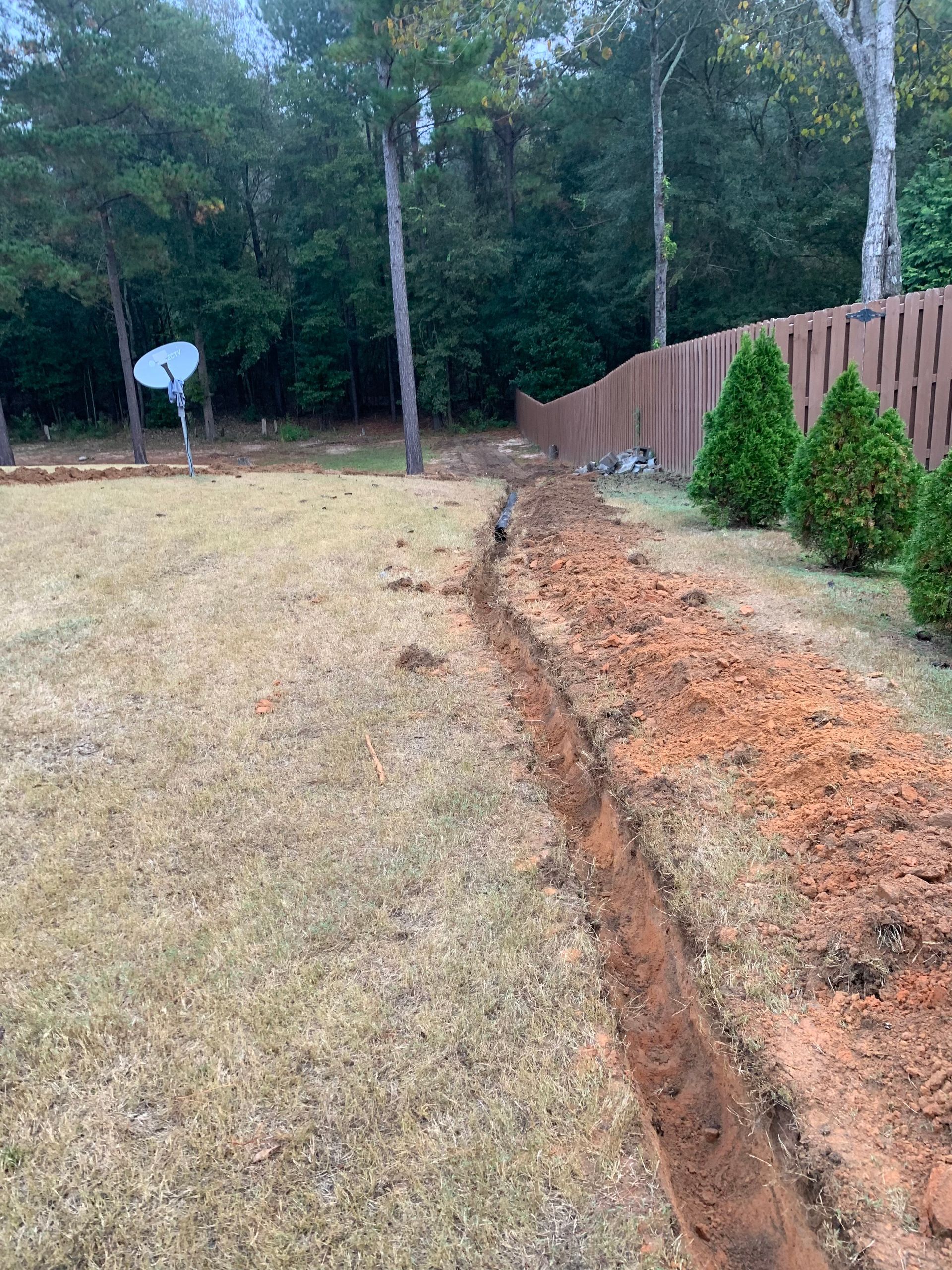 Narrow dirt trench along a yard beside a wooden fence and shrubs, with trees in the background.
