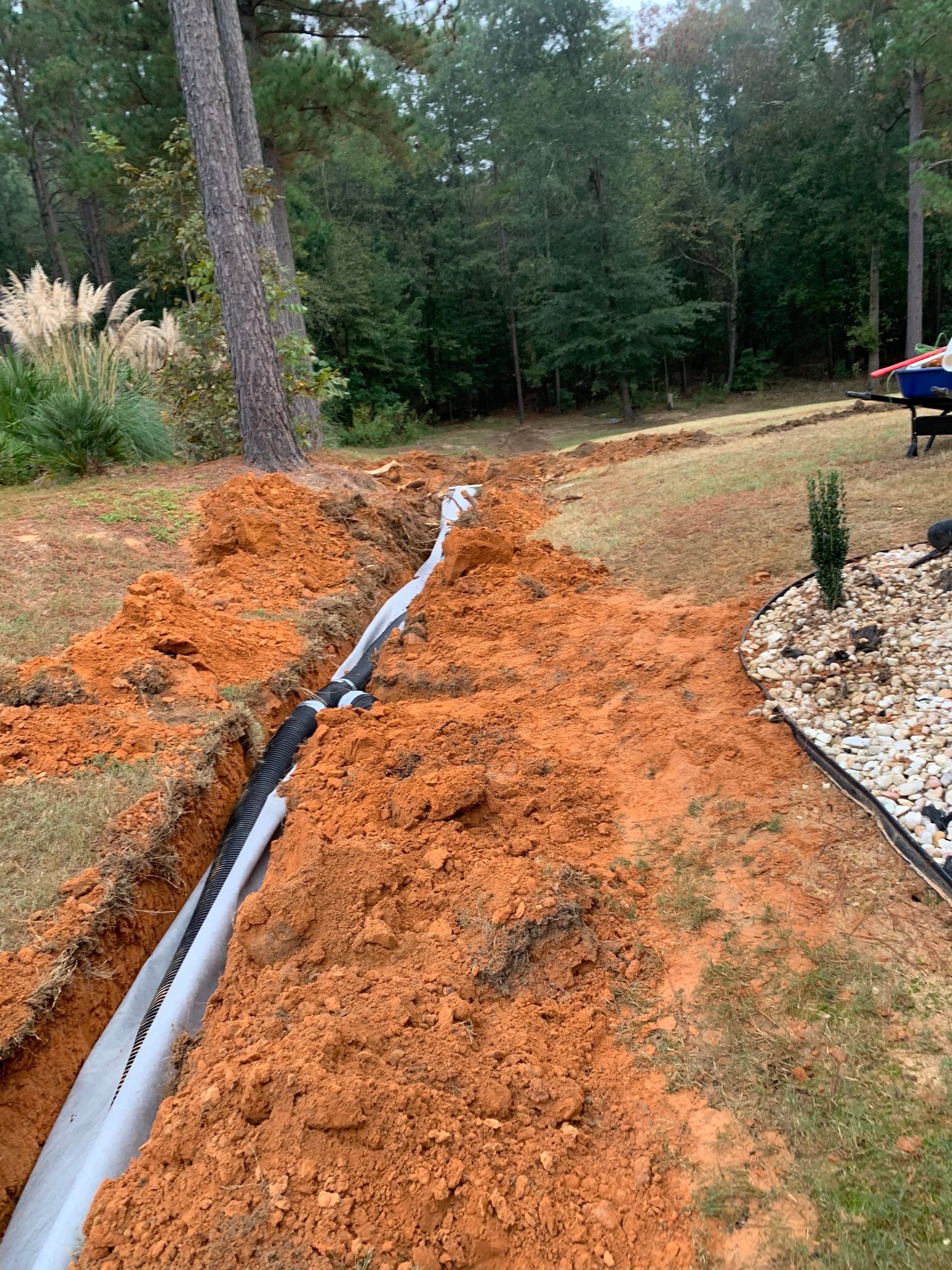 Orange dirt trench with a gray pipe running through a yard beside a rock border and trees