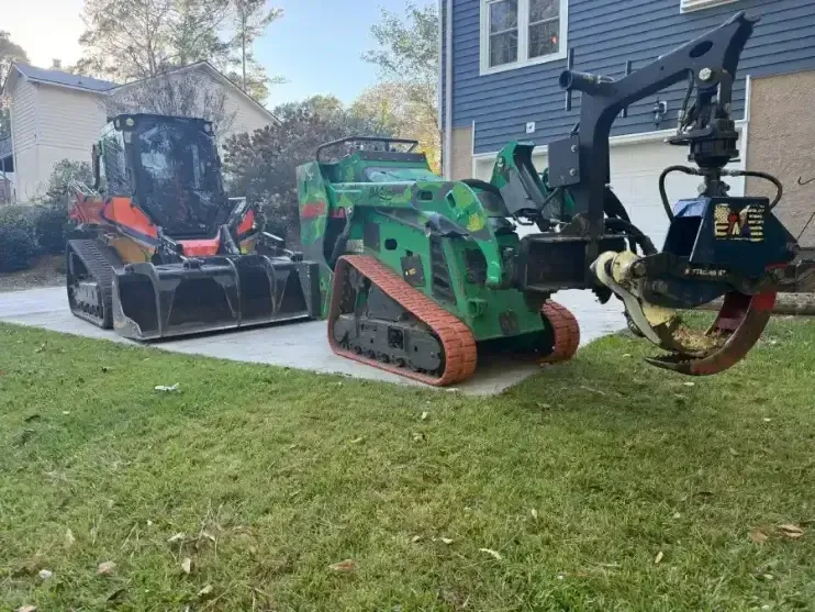 Two pieces of heavy equipment, an orange skid steer and a green forestry loader, parked on a suburban driveway.
