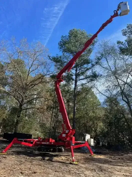A red track-mounted spider lift with an extended articulated boom and basket set up in a wooded area under a blue sky.