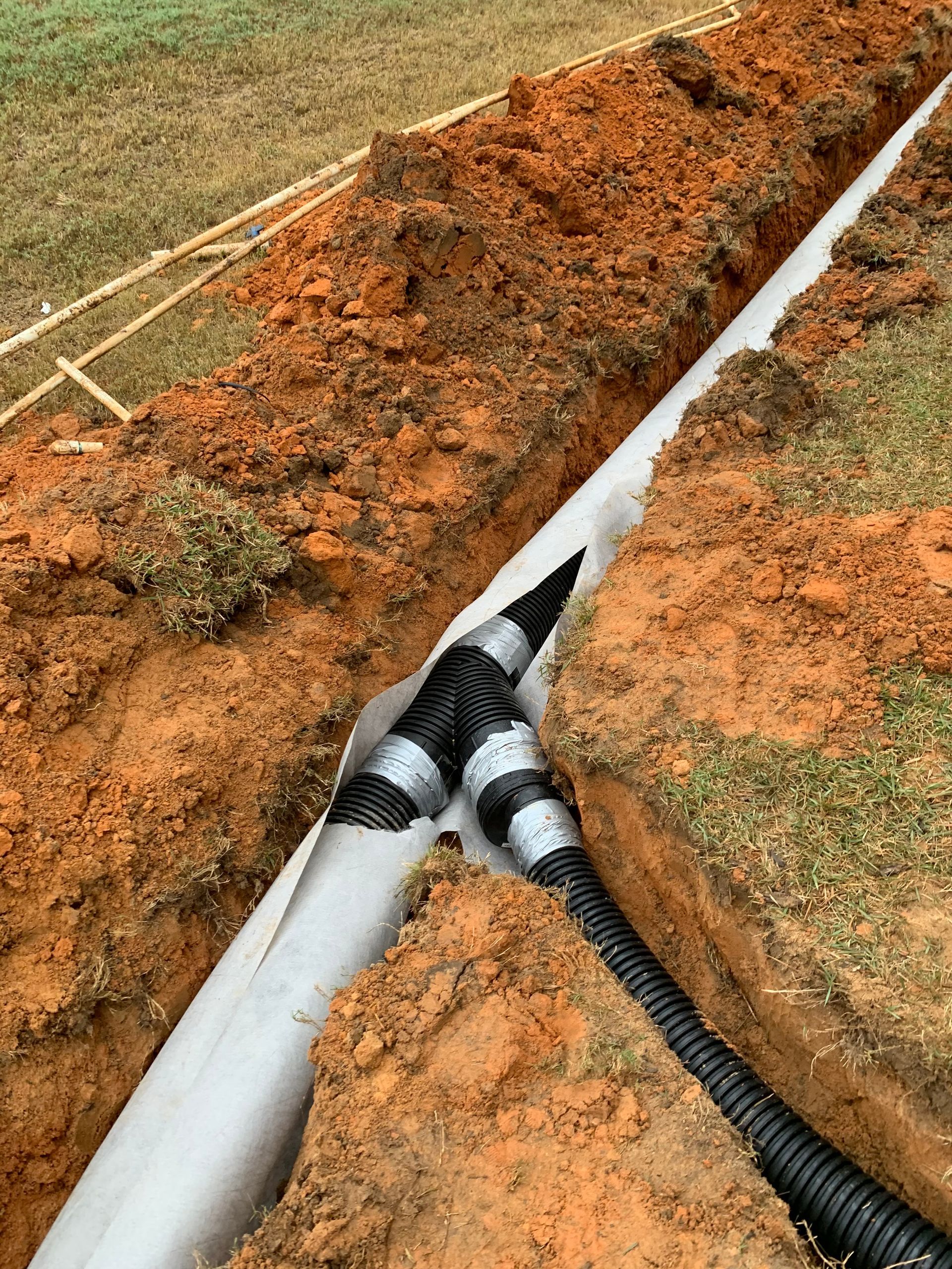 Utility trench with white pipe and black cable laid in red dirt grass along the edge
