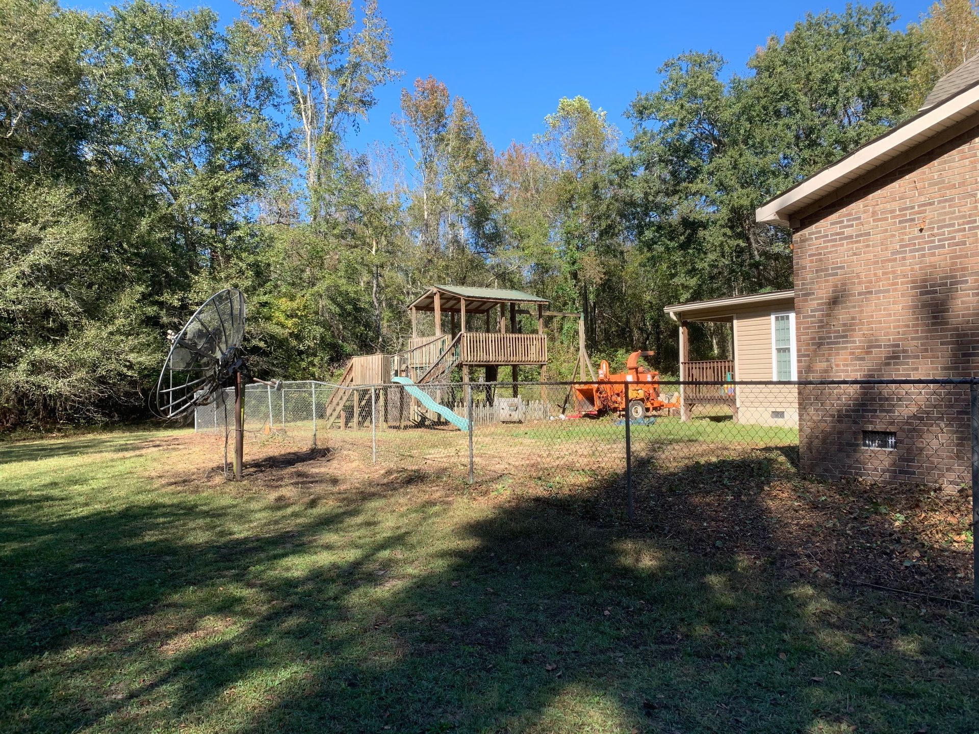 Backyard with a playset, orange yard equipment, chain-link fence, trees, and a brick house under clear sky