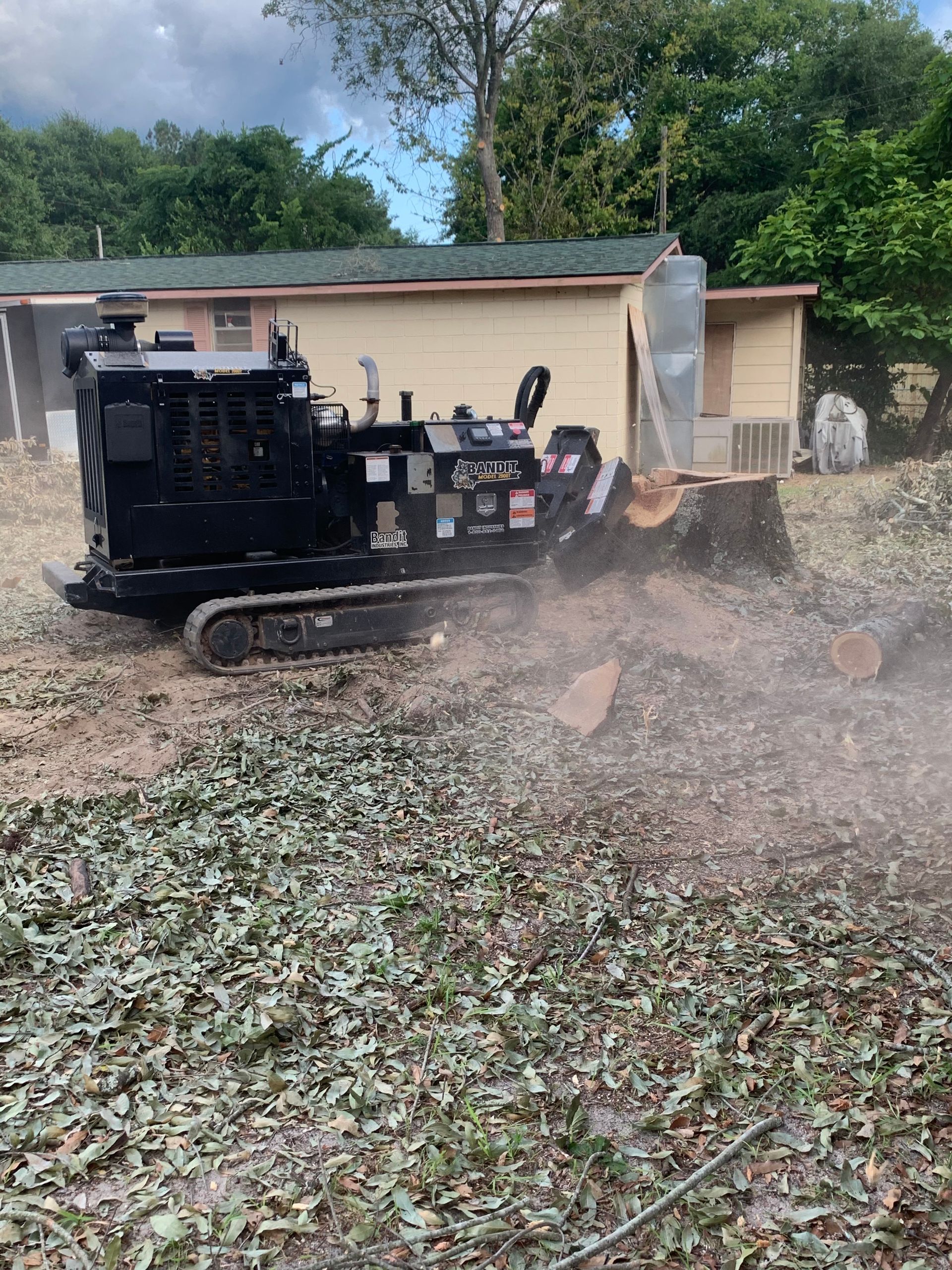 Black tracked stump grinder kicking up wood chips in a yard beside a white garage.