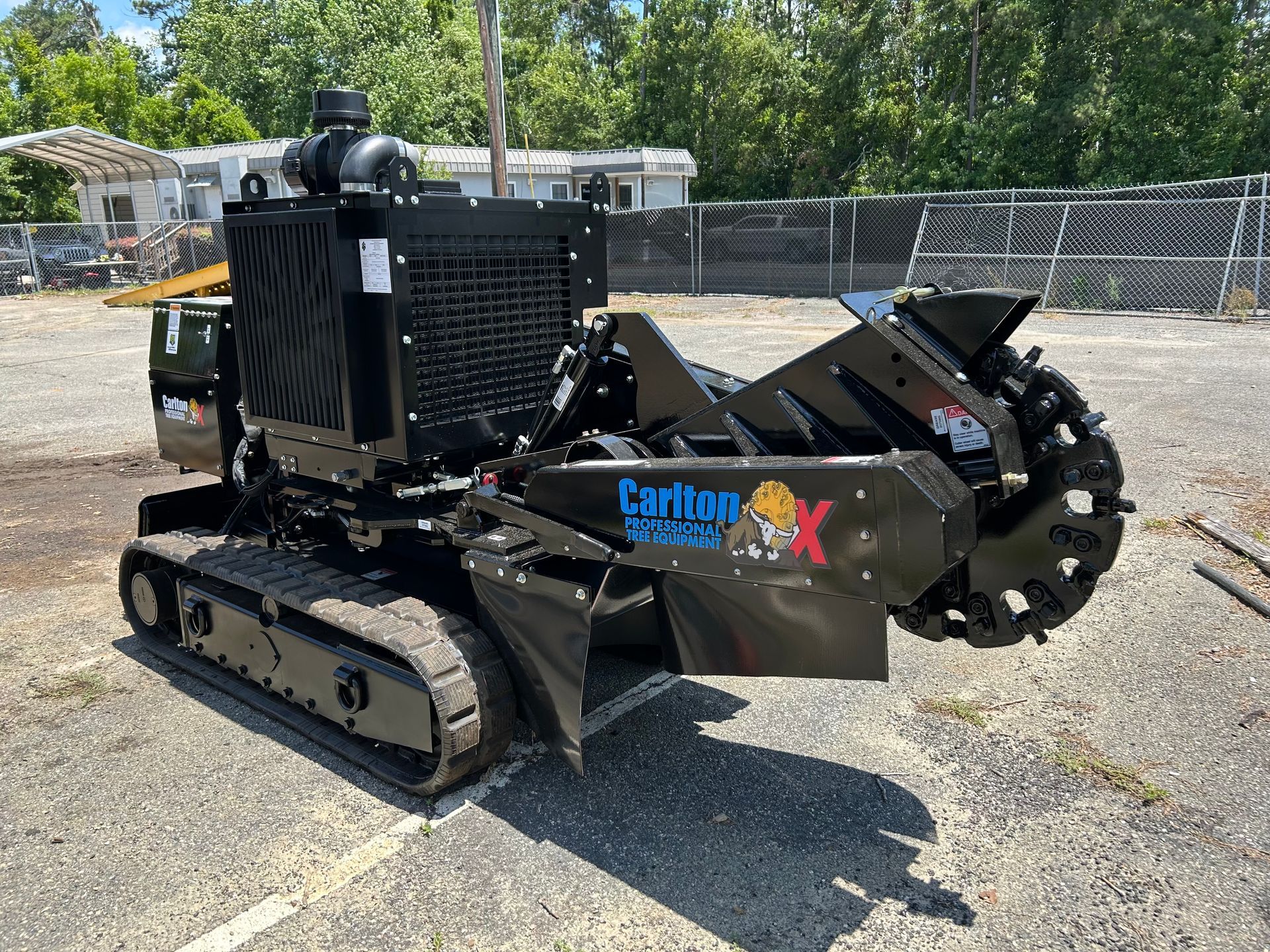 Black tracked stump grinder parked on gravel lot with trees and fencing in the background