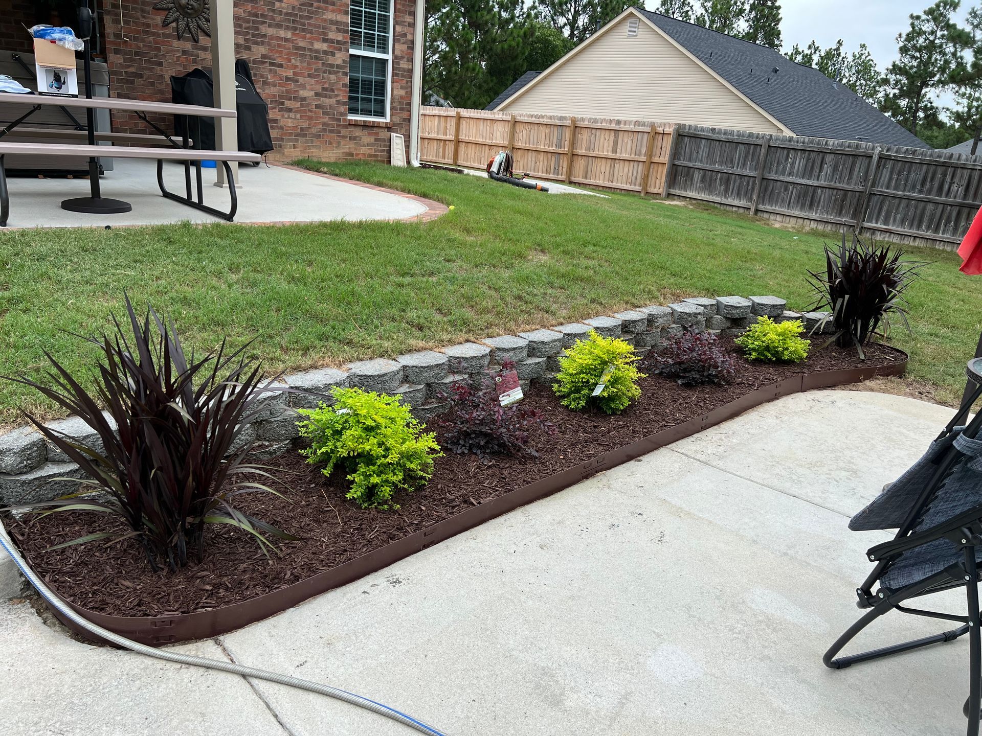 Landscaped backyard with mulch bed, small shrubs, and a concrete patio beside a house and fence.