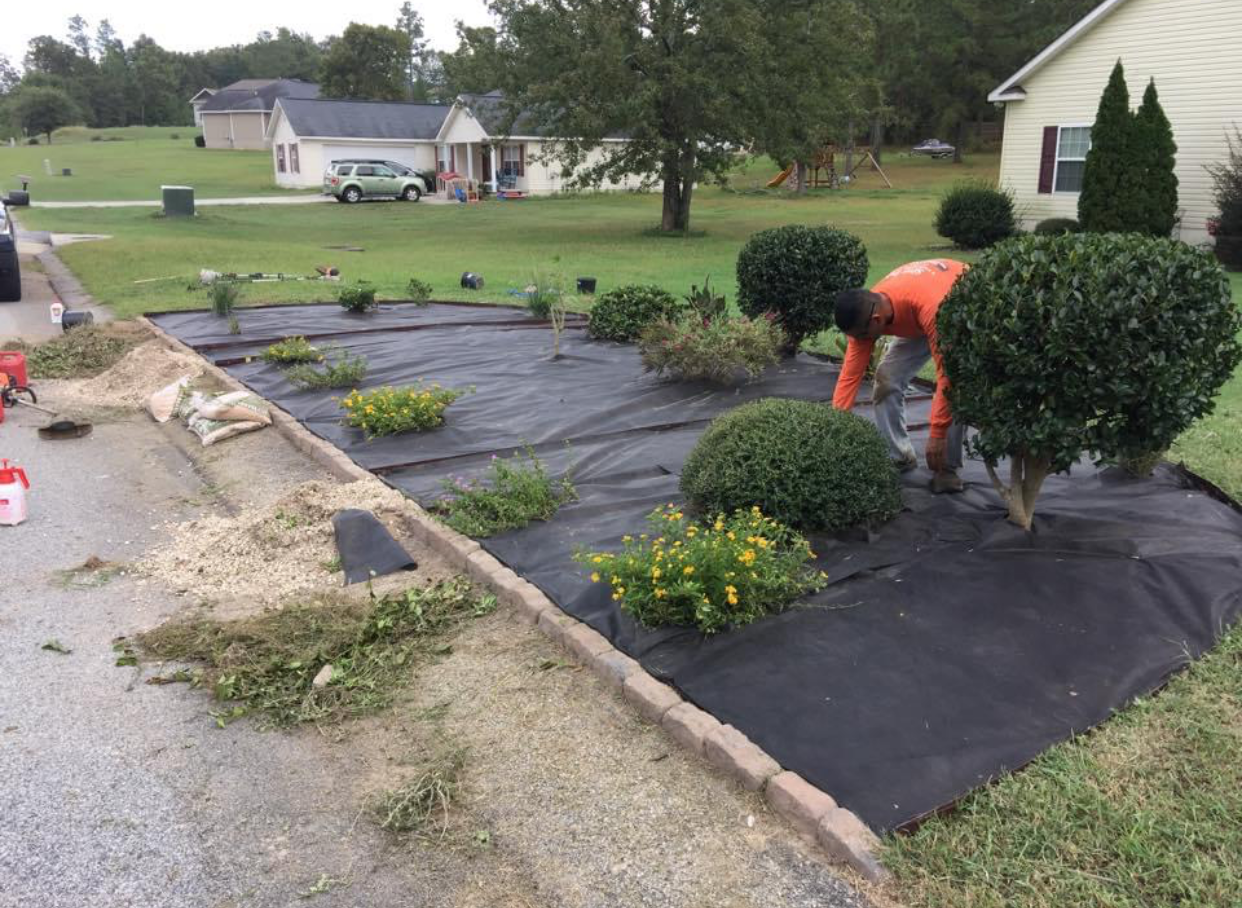 Person laying black landscape fabric in a garden bed beside a curb, with shrubs and a house in the background