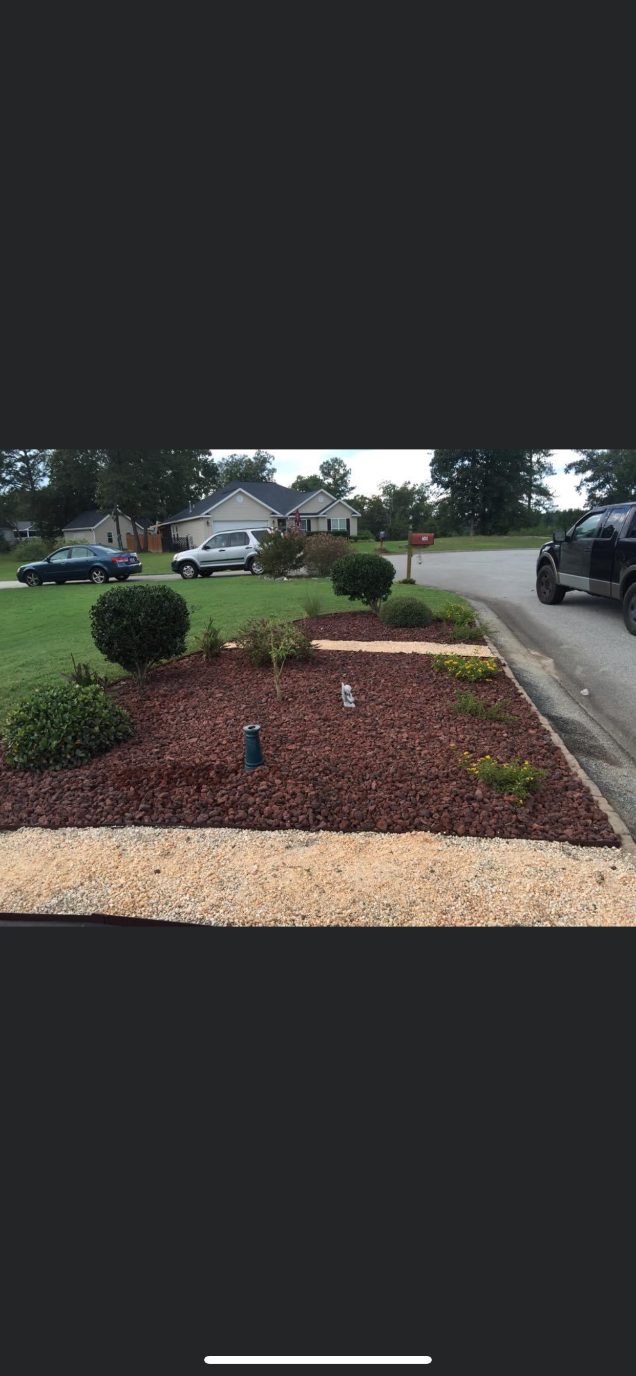 Mulched front yard garden bed with small shrubs and a gravel walkway beside a street and parked cars