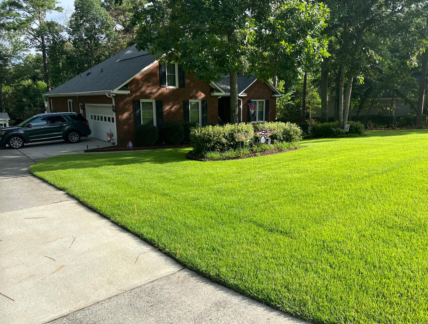 Suburban brick house with a black car in the driveway and a green lawn under trees