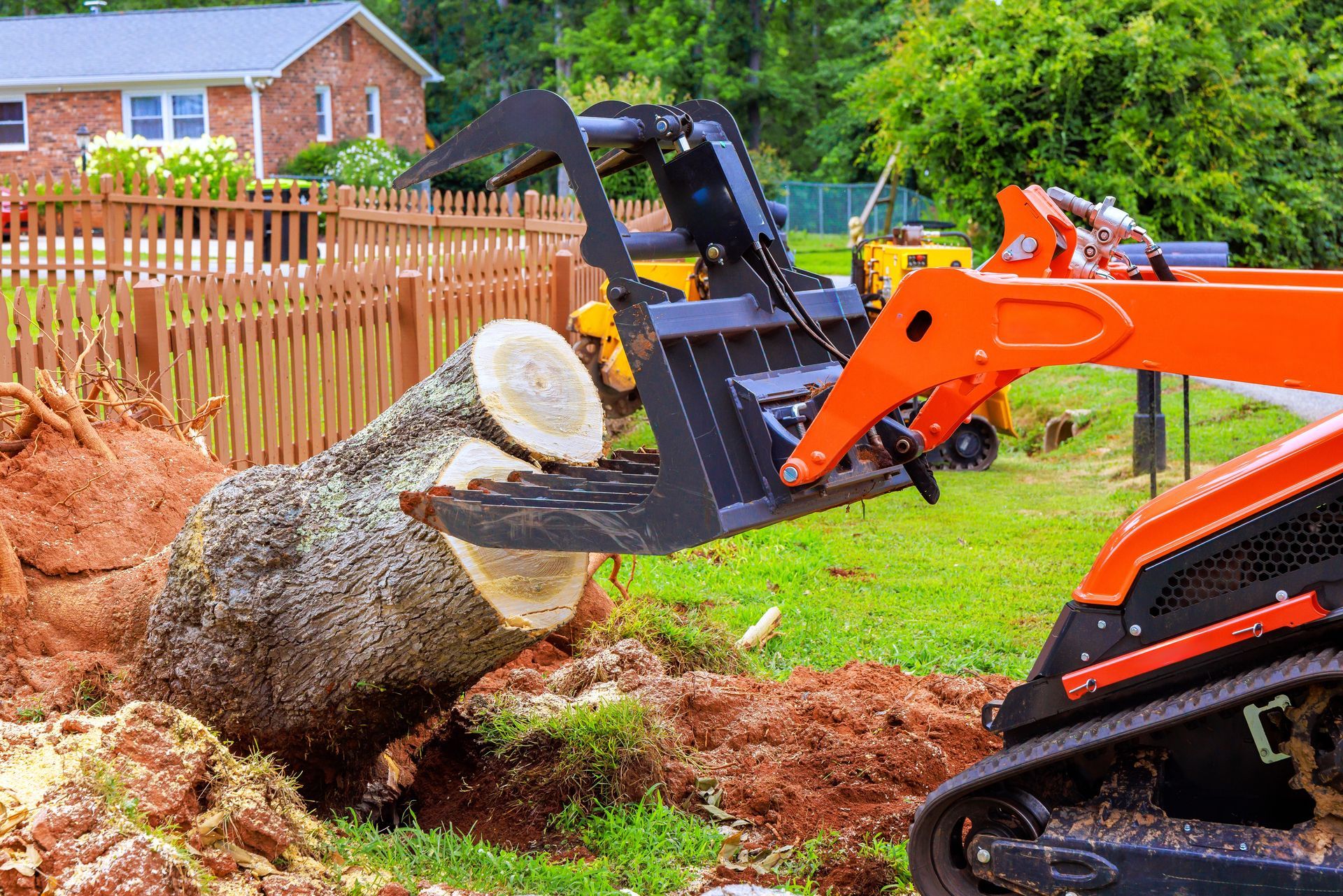 A bright orange skid steer with a black hydraulic claw attachment lifting a large tree stump in a grassy backyard.