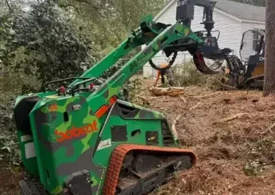 Green and camouflage Bobcat mini track loader with a grapple attachment clearing brush in a residential yard.