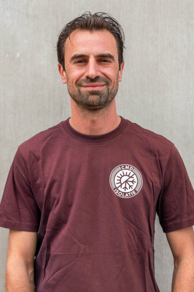 Man in een kastanjebruin t-shirt met een logo glimlacht tegen een grijze muur.