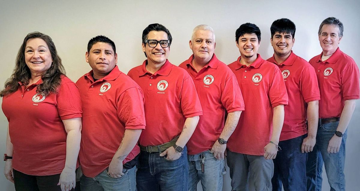 Group of seven people in red polo shirts, likely a team, standing indoors against a plain wall, smiling.